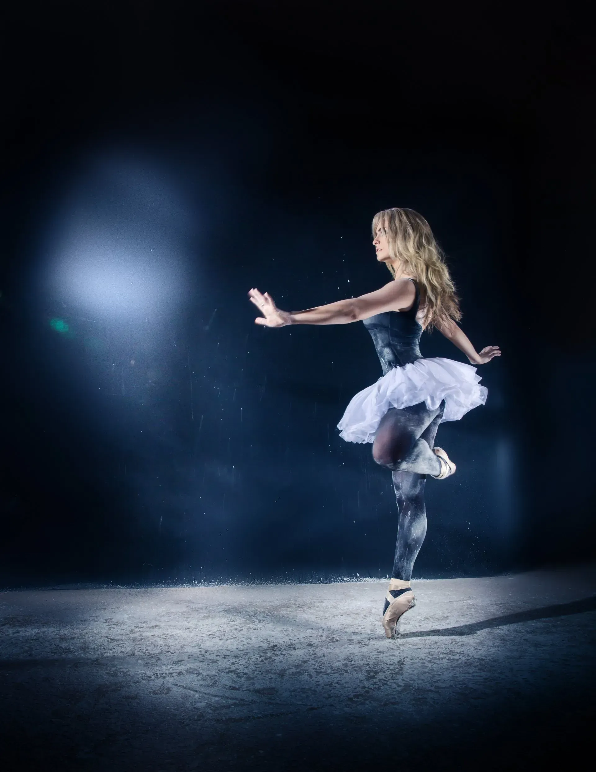 Ballerina in a white tutu dancing on pointe under blue spotlight on a dark stage