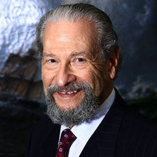 Smiling man in a dark suit and red tie, posed against a blurred mountain backdrop.