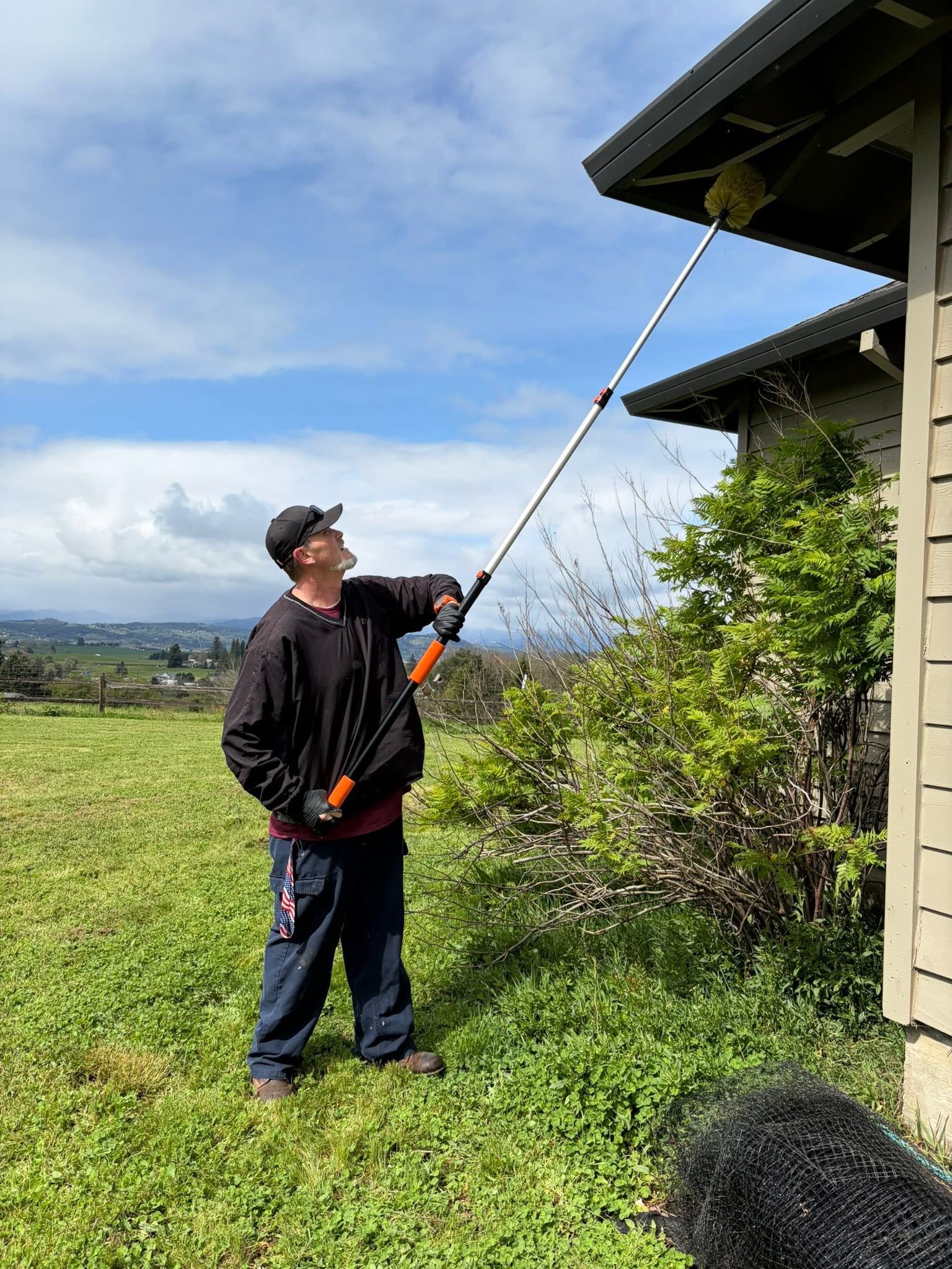 Person using a long pole to inspect the roof edge of a house on a grassy property.