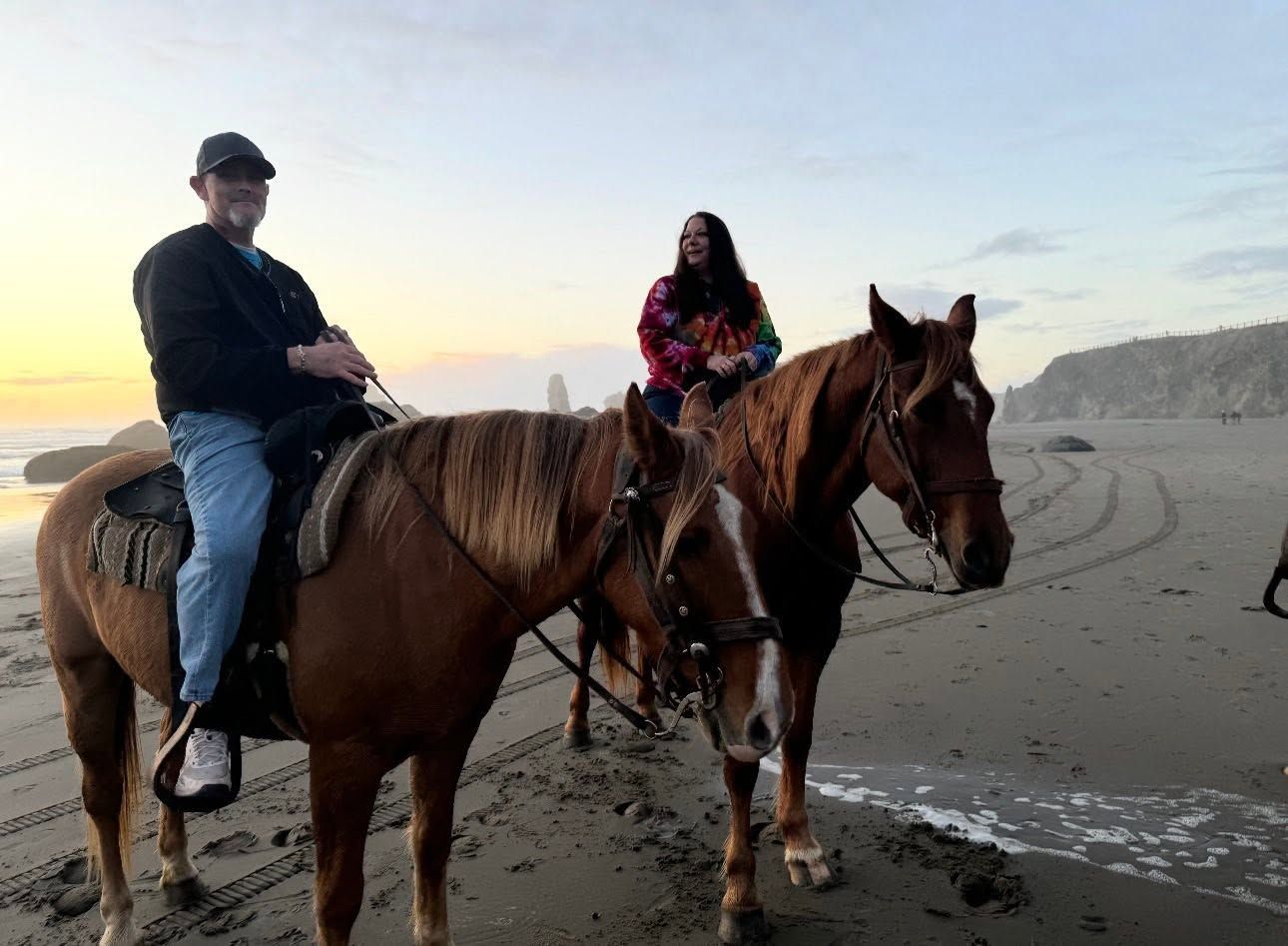 Two people ride horses on a beach at sunset, with wet sand and ocean waves nearby.