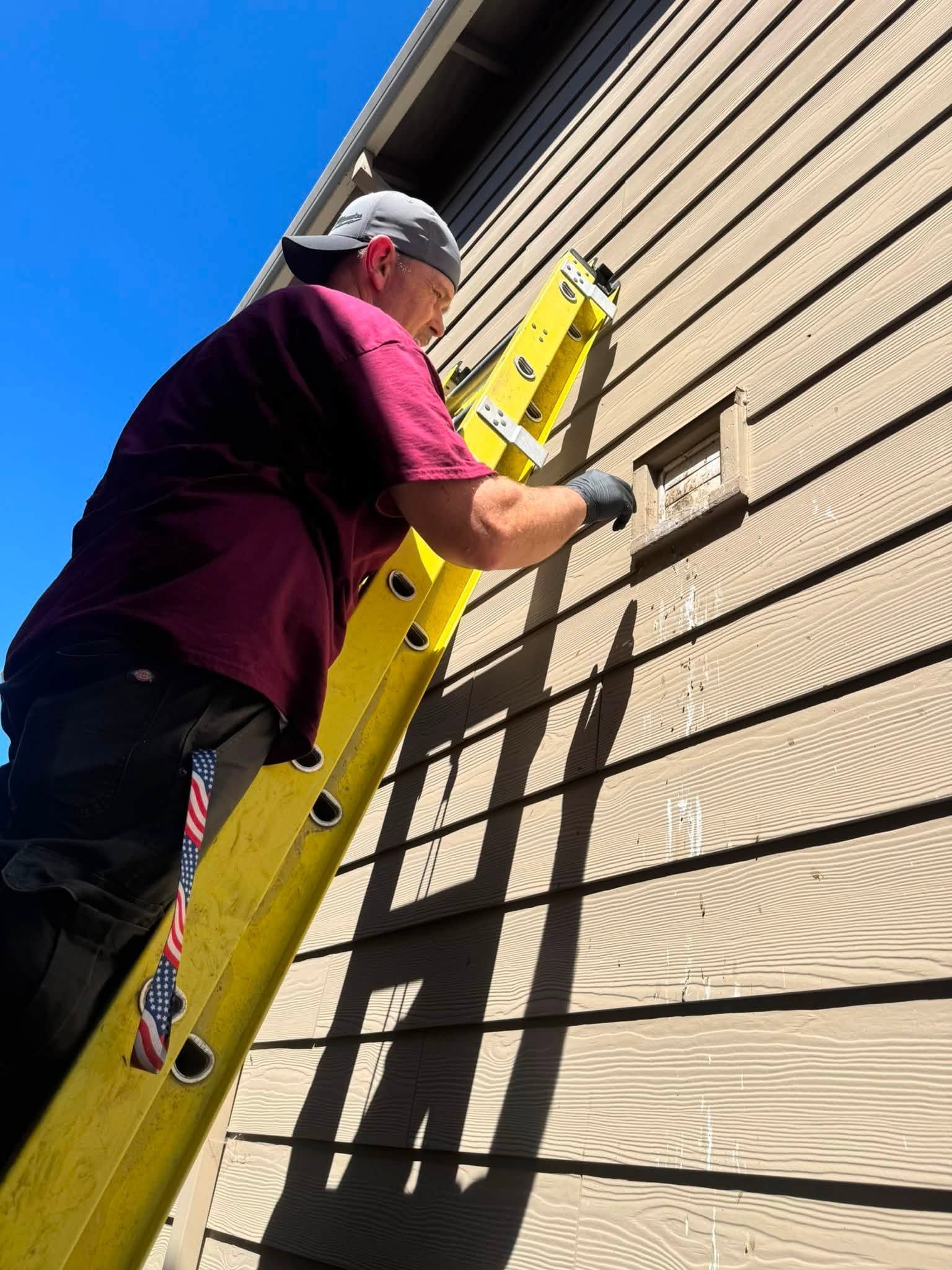 Worker on a yellow ladder painting a tan house exterior under a clear blue sky