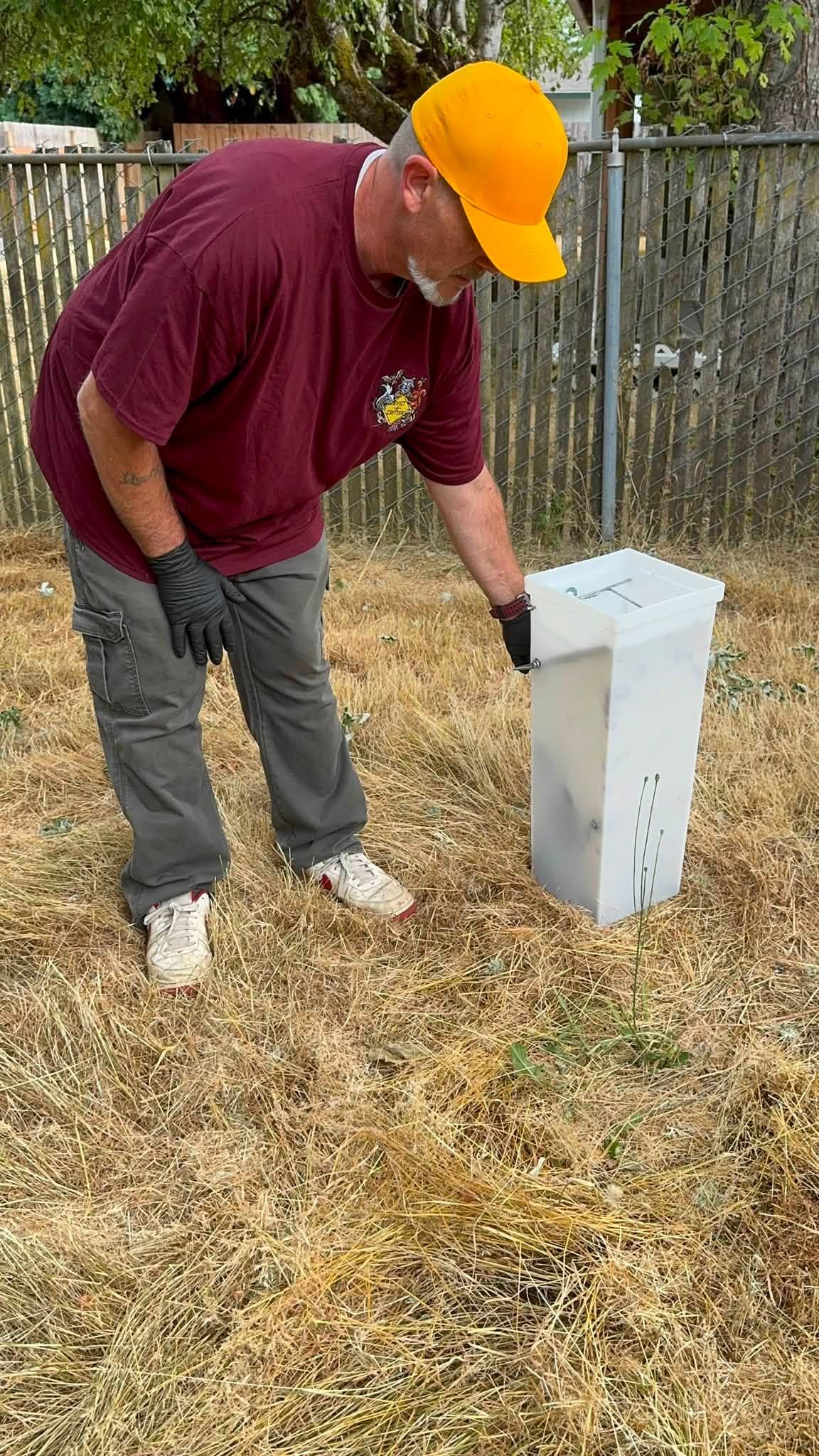 Man in orange hard hat points at a white utility box in a dry yard