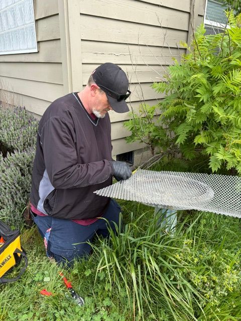 Person kneeling outdoors, using a tool to cut a metal mesh panel beside a house and shrubs