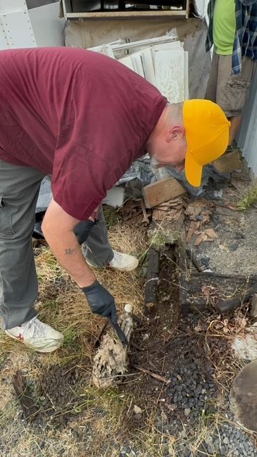Person in a yellow cap bends over clearing debris beside a stone wall outdoors