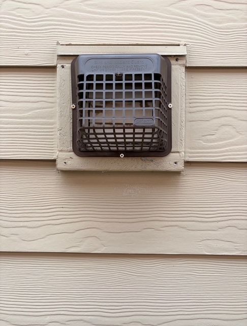 Exterior wall vent with a black protective cage on beige siding