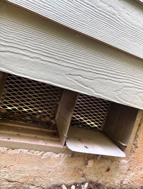 Exterior wall vent covered with white siding and metal mesh, above a stone foundation