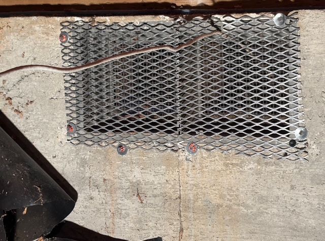 Metal mesh vent mounted on a rough concrete wall with exposed wiring above it