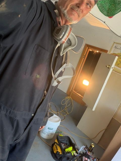Person in a dark hoodie and respirator works in a room with tools, wires, and a lit lamp.