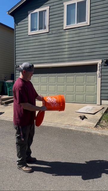 Person holding an orange bucket in front of a garage in a residential driveway.