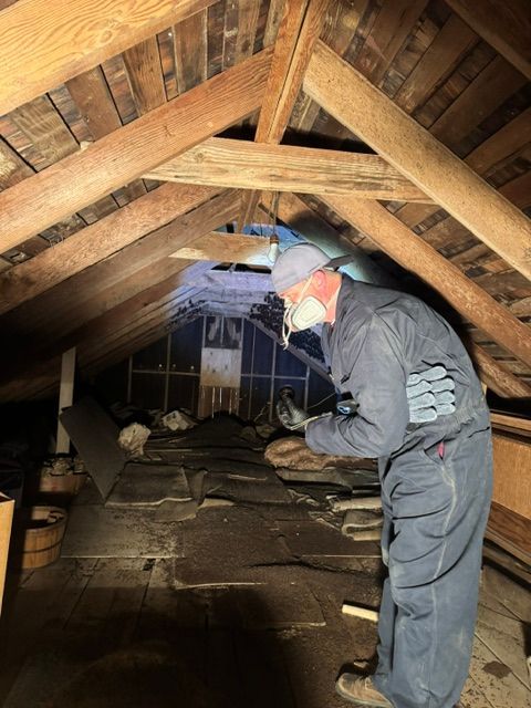 Person in attic inspecting insulation with a flashlight amid exposed wooden beams.