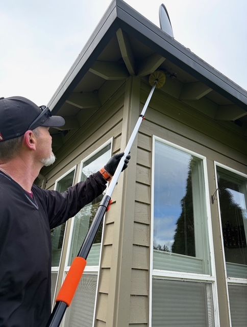 Person uses a long tool to clean a house roof edge and siding near a window.