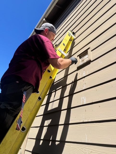 Person on a yellow ladder painting beige house siding under a blue sky