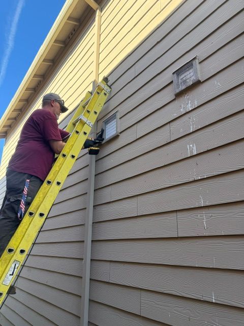 Person on a yellow ladder repairing siding on a beige house under a blue sky