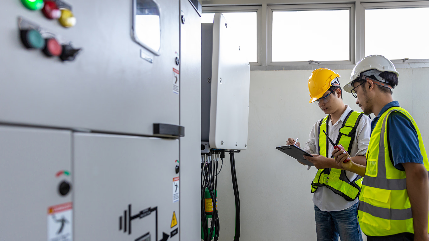 Two workers in hard hats inspect electrical equipment indoors, one holding a clipboard.