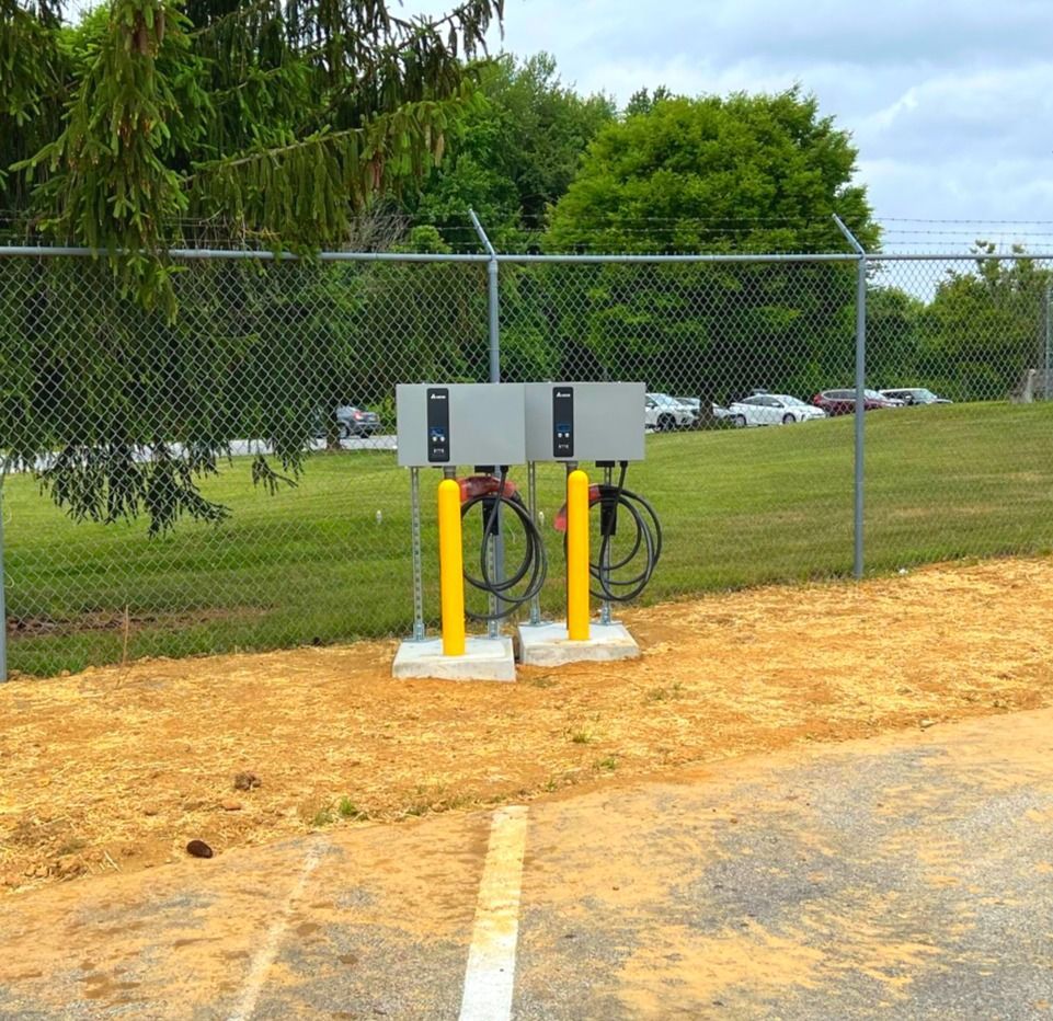 Chain-link fence with two gray electrical boxes on yellow posts beside a parking lot and trees