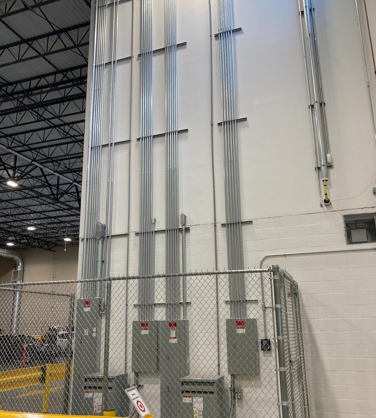 Industrial electrical panels and conduit pipes mounted on a warehouse wall behind a chain-link fence.