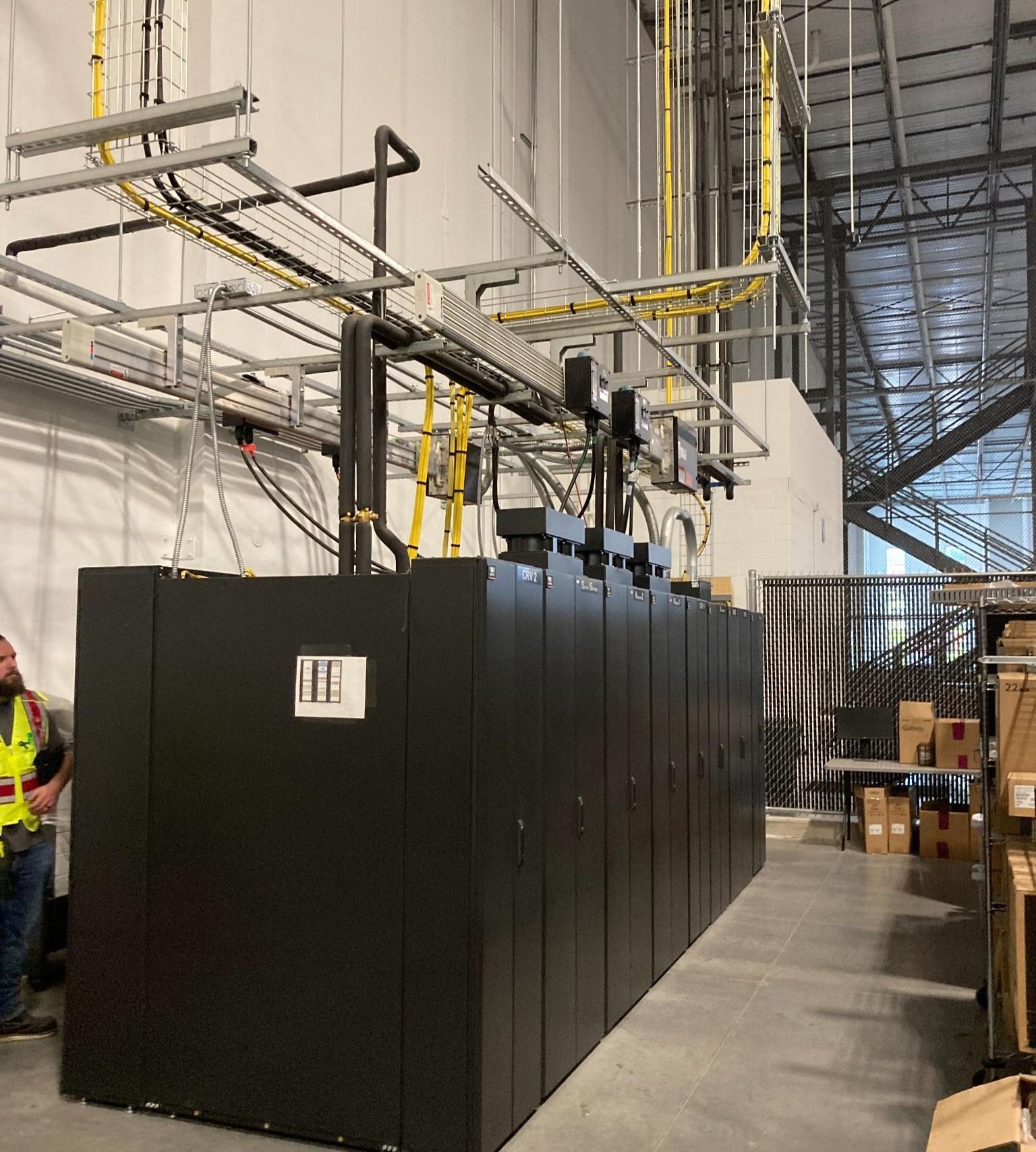 Large black industrial equipment cabinets in a warehouse aisle with overhead cable trays and shelving