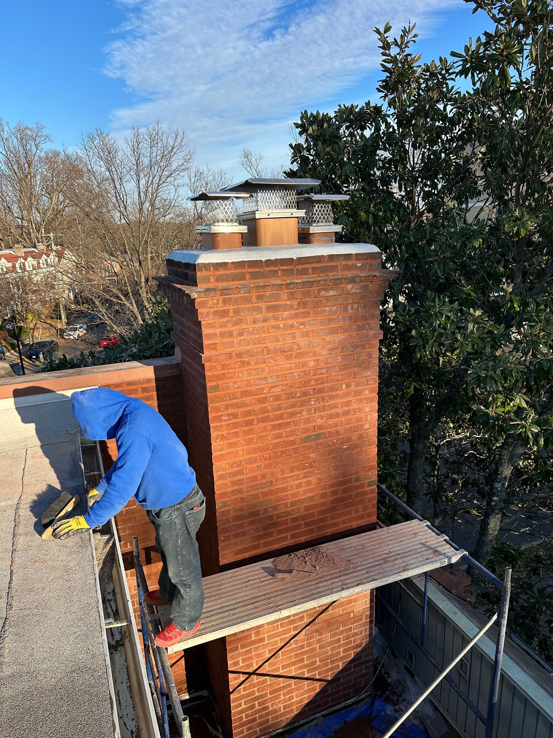 A man in a blue jacket is working on a brick chimney on a roof.