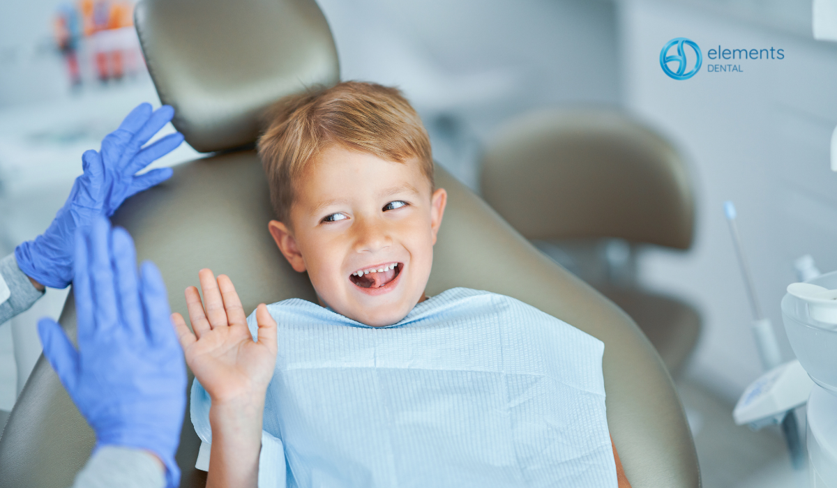 Smiling child in dental chair with blue bib, while dentist in blue gloves waves nearby