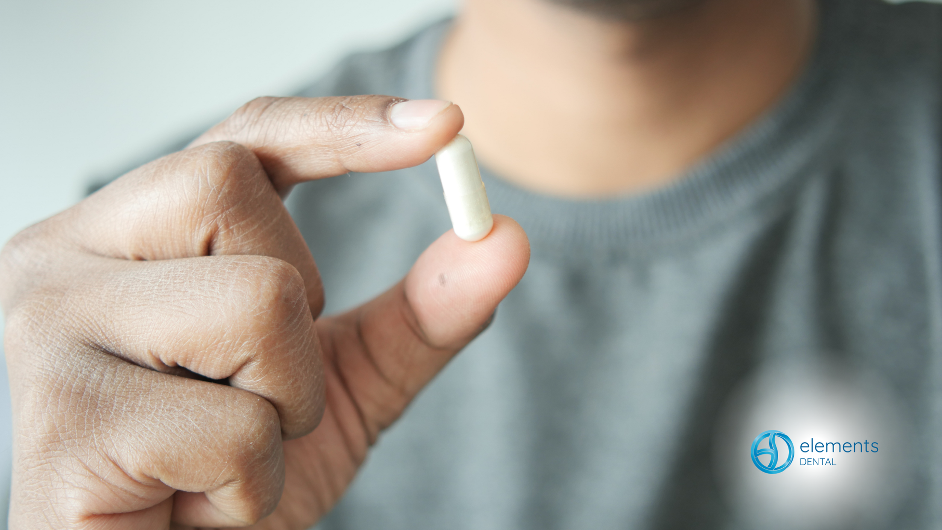 Close-up of a hand pinching a tiny white pill between thumb and forefinger, with a blurred gray shirt background