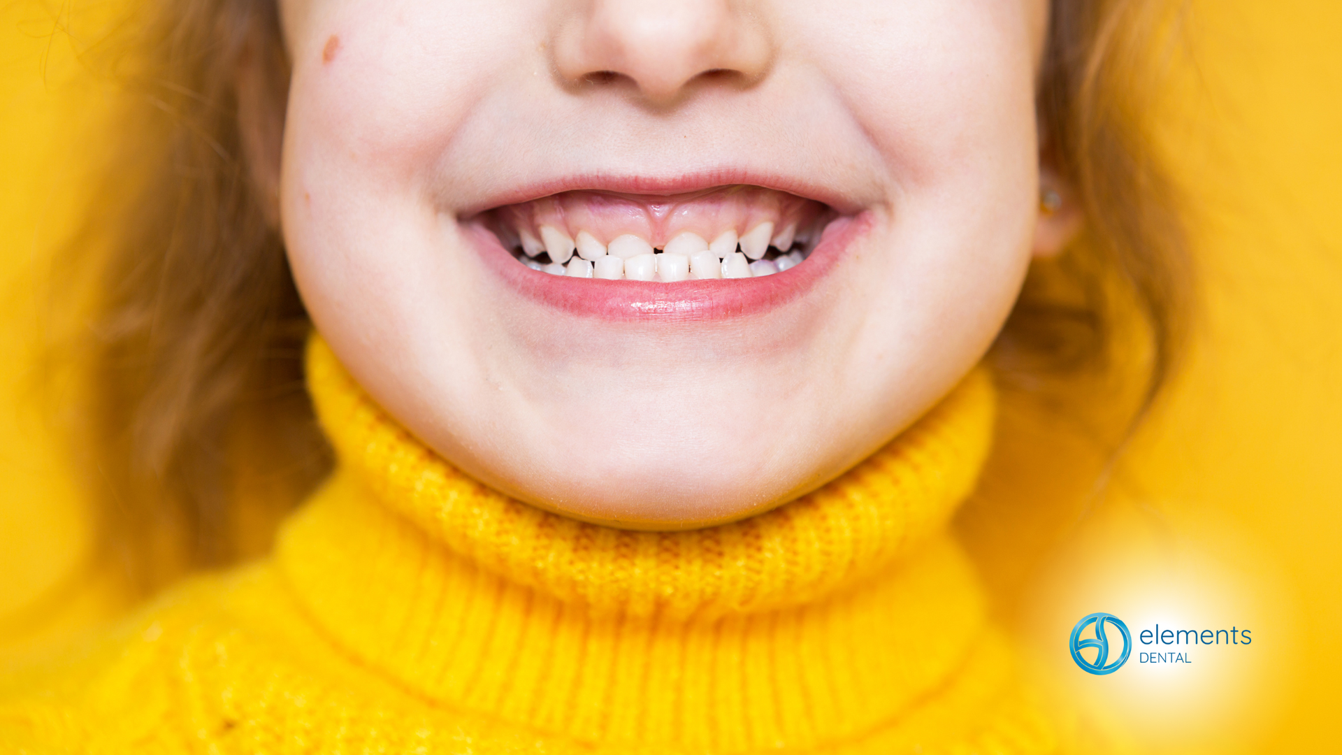 A little girl wearing a yellow sweater is smiling and showing her teeth.