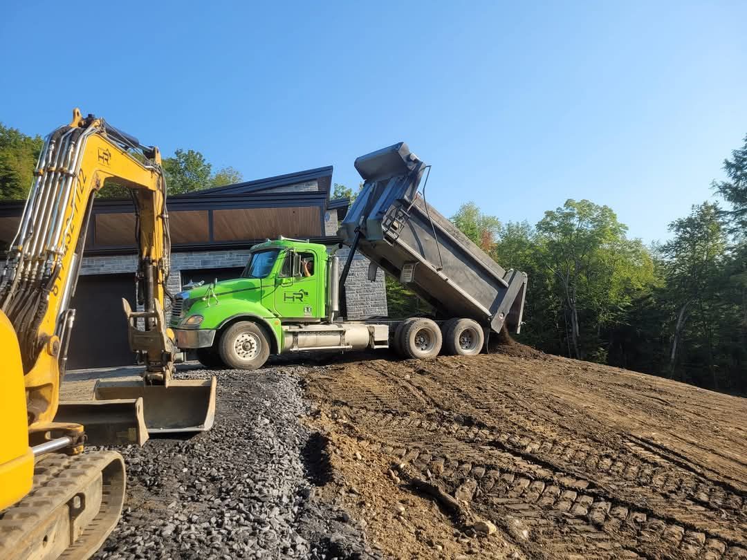 Un camion-benne vert décharge de la terre, une excavatrice jaune se trouve à proximité, en extérieur sous un ciel bleu.