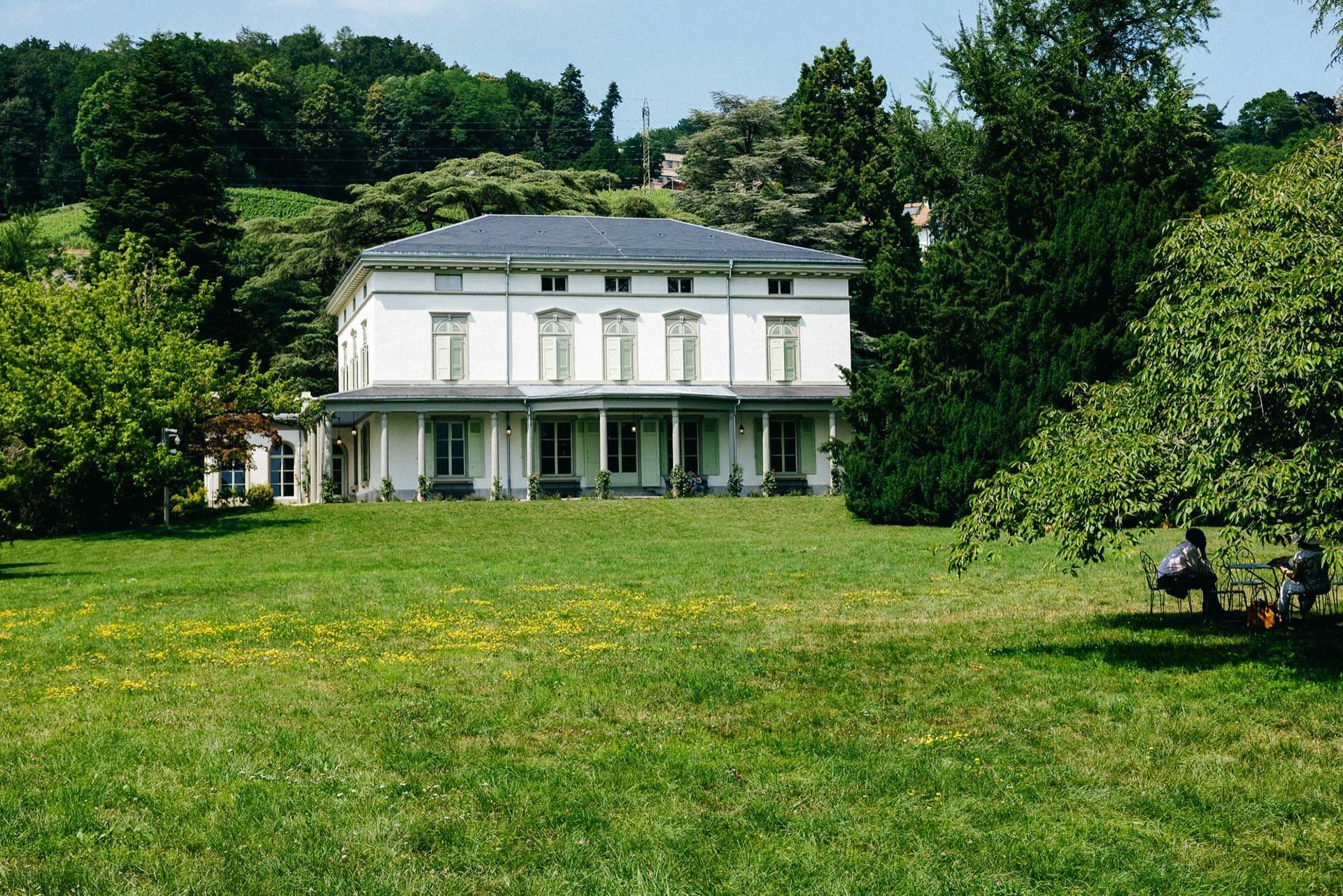 Bâtiment blanc avec une longue véranda, situé dans une cour herbeuse, avec des arbres en arrière-plan. Deux personnes sont assises sur un banc.