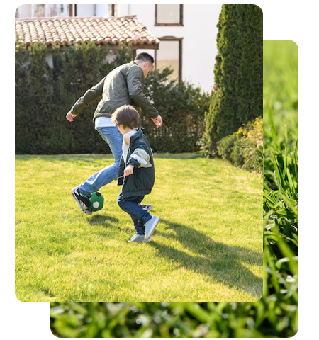 Un homme et un enfant jouent au football dans un jardin ensoleillé, avec une maison et des plantes vertes en arrière-plan.