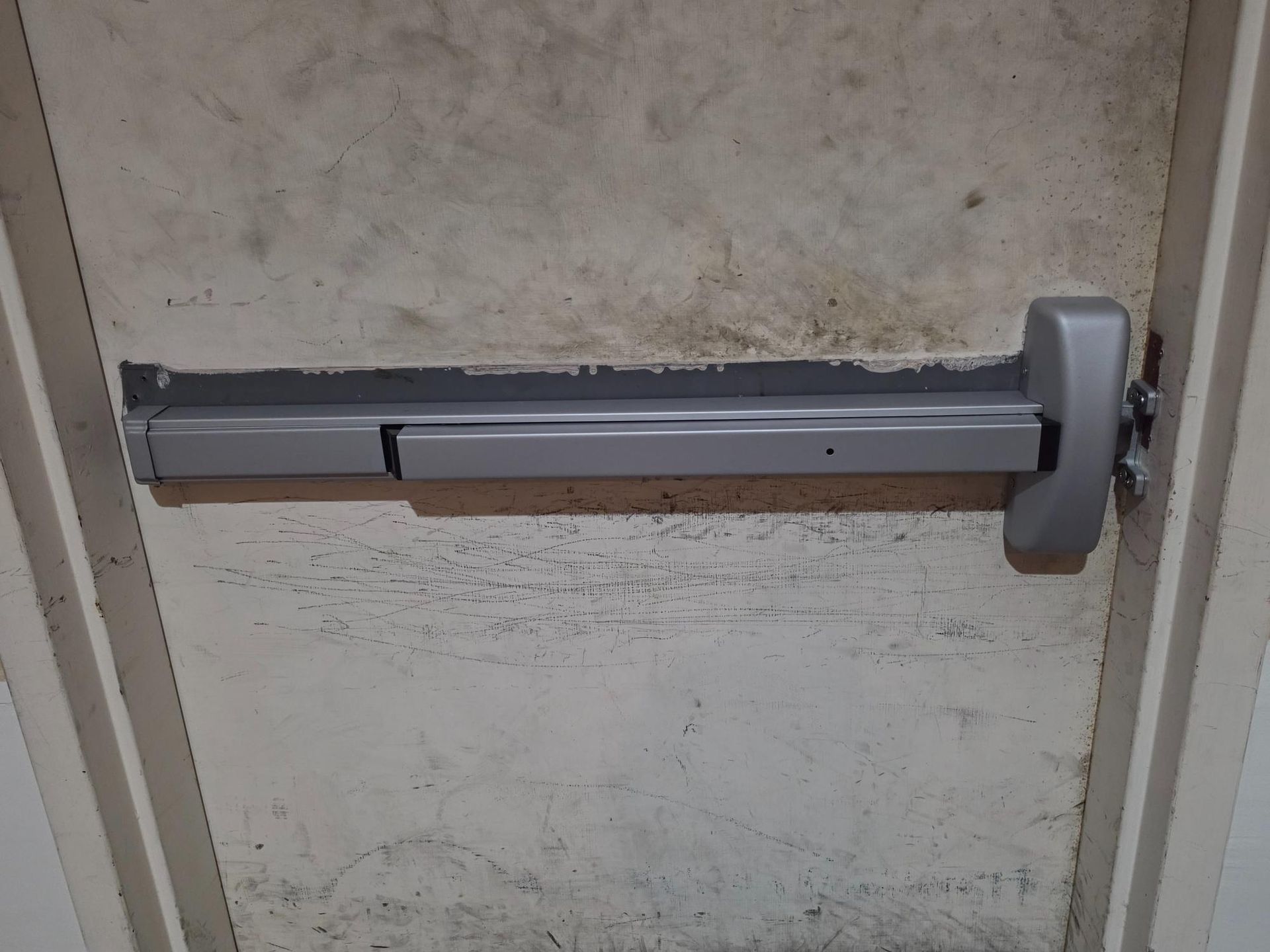 A light-colored horizontal panic bar exit device mounted on a plain, weathered interior door.