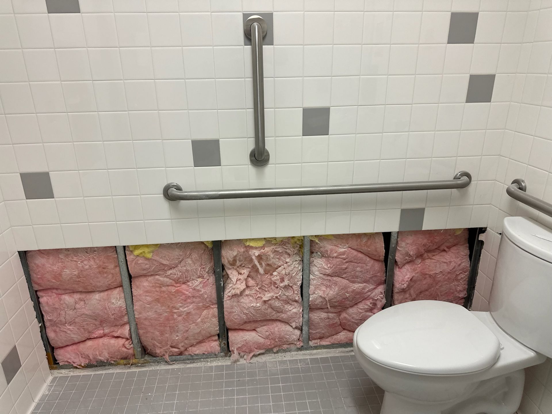 A damaged restroom stall wall with exposed pink insulation beneath tiled grab bars and a toilet.