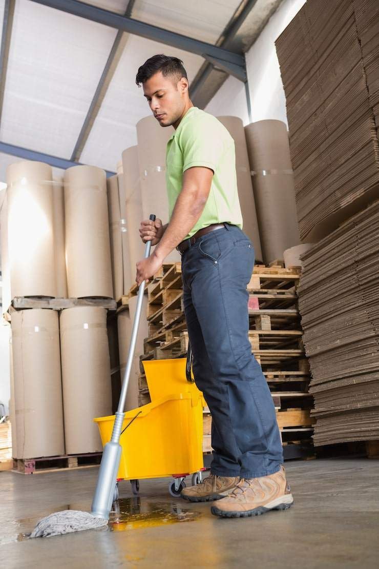 A worker in a lime-green shirt mops a spill on a concrete warehouse floor near stacked rolls and wooden pallets.