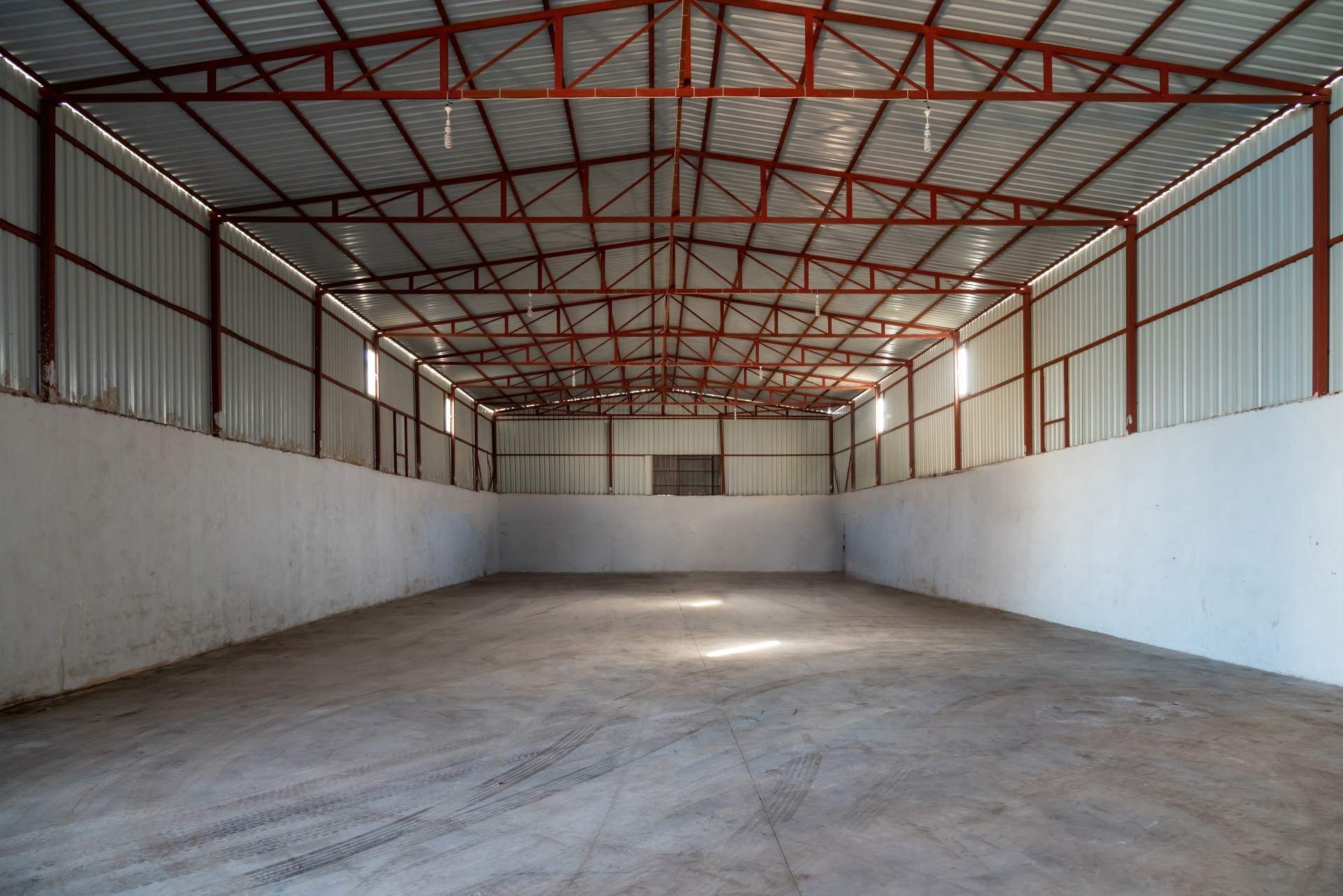 A spacious, empty industrial warehouse with white concrete walls, a polished gray floor, and a red metal truss roof.