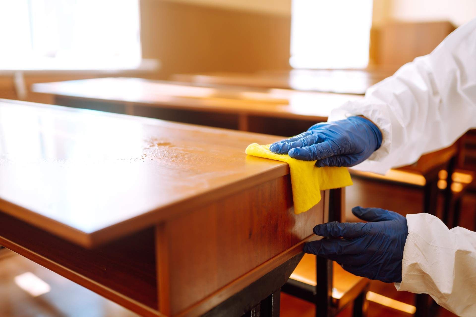 A person in a white protective suit and blue gloves wipes a wooden school desk with a yellow cloth.