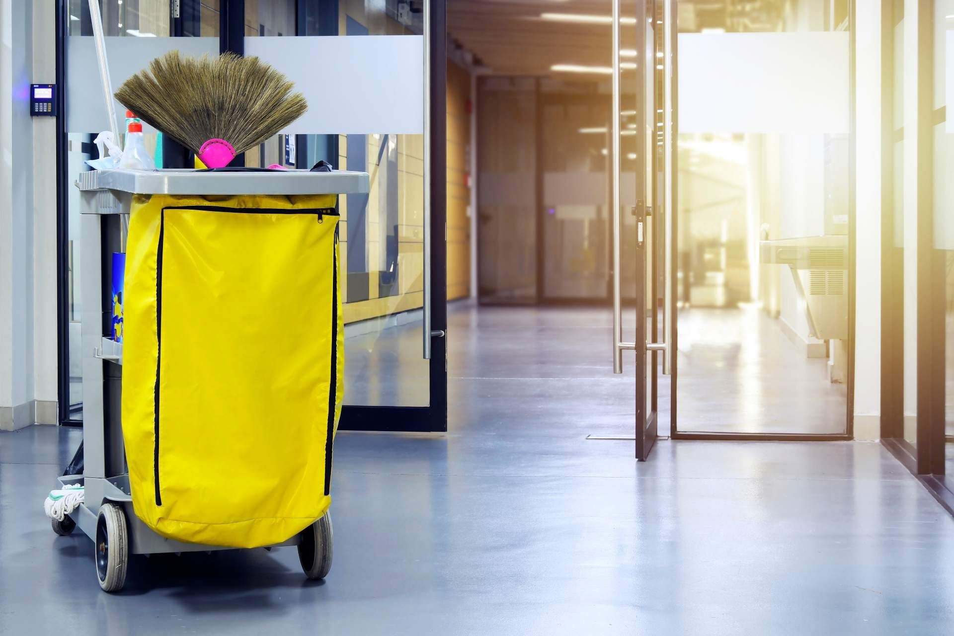 A yellow-bagged janitorial cleaning cart stands on a polished floor in a bright, modern office hallway.