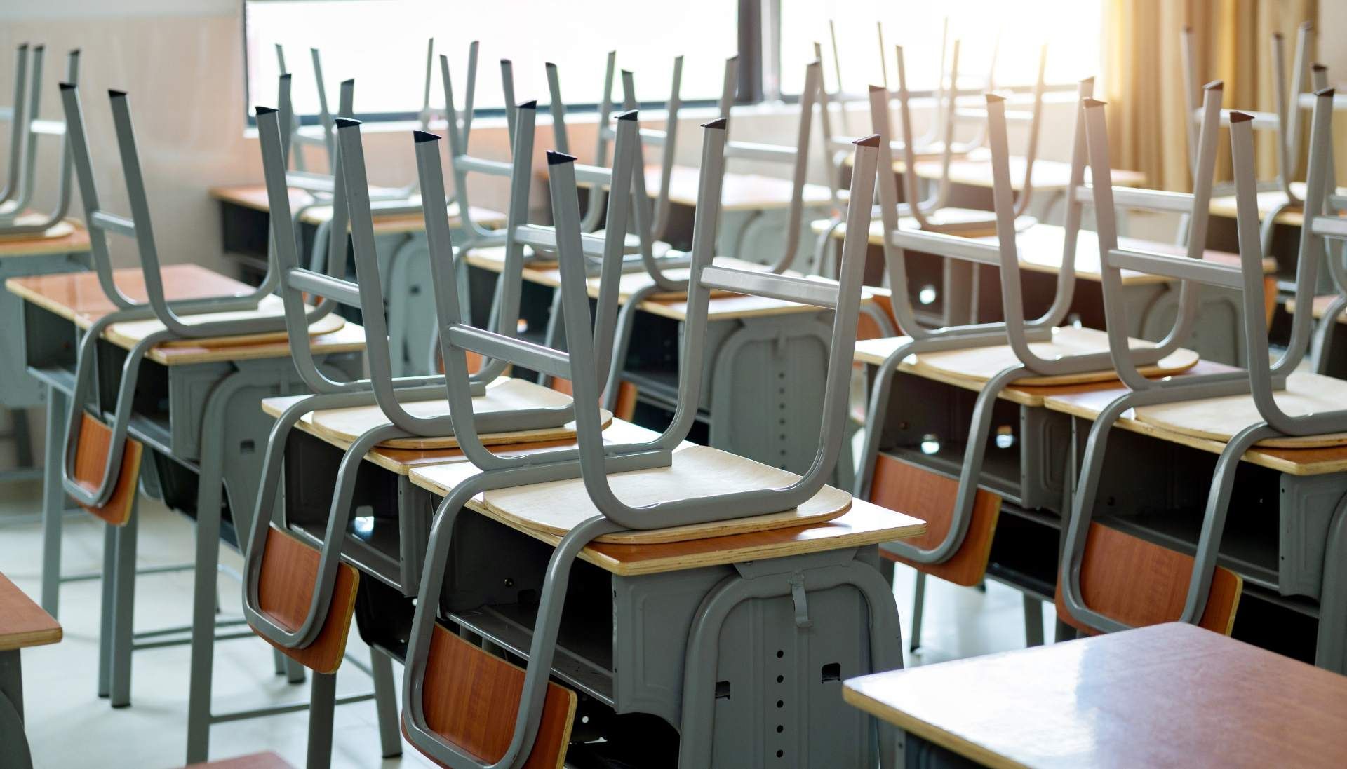 Empty school desks with chairs stacked on top, positioned in rows inside a brightly lit classroom.