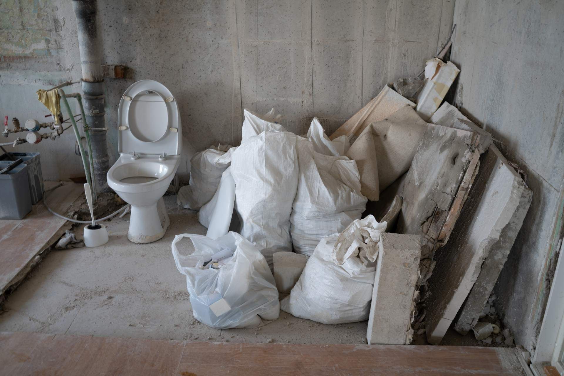 A white toilet stands in a room undergoing construction, next to piles of white debris bags and concrete slabs.