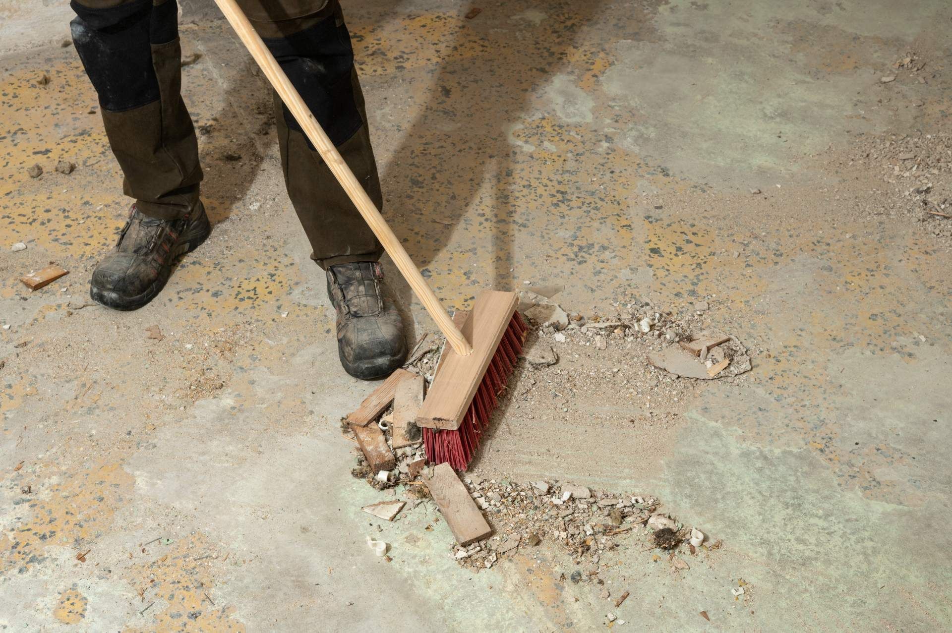 A person wearing sturdy work boots uses a push broom to clear construction debris and dust from a concrete floor.