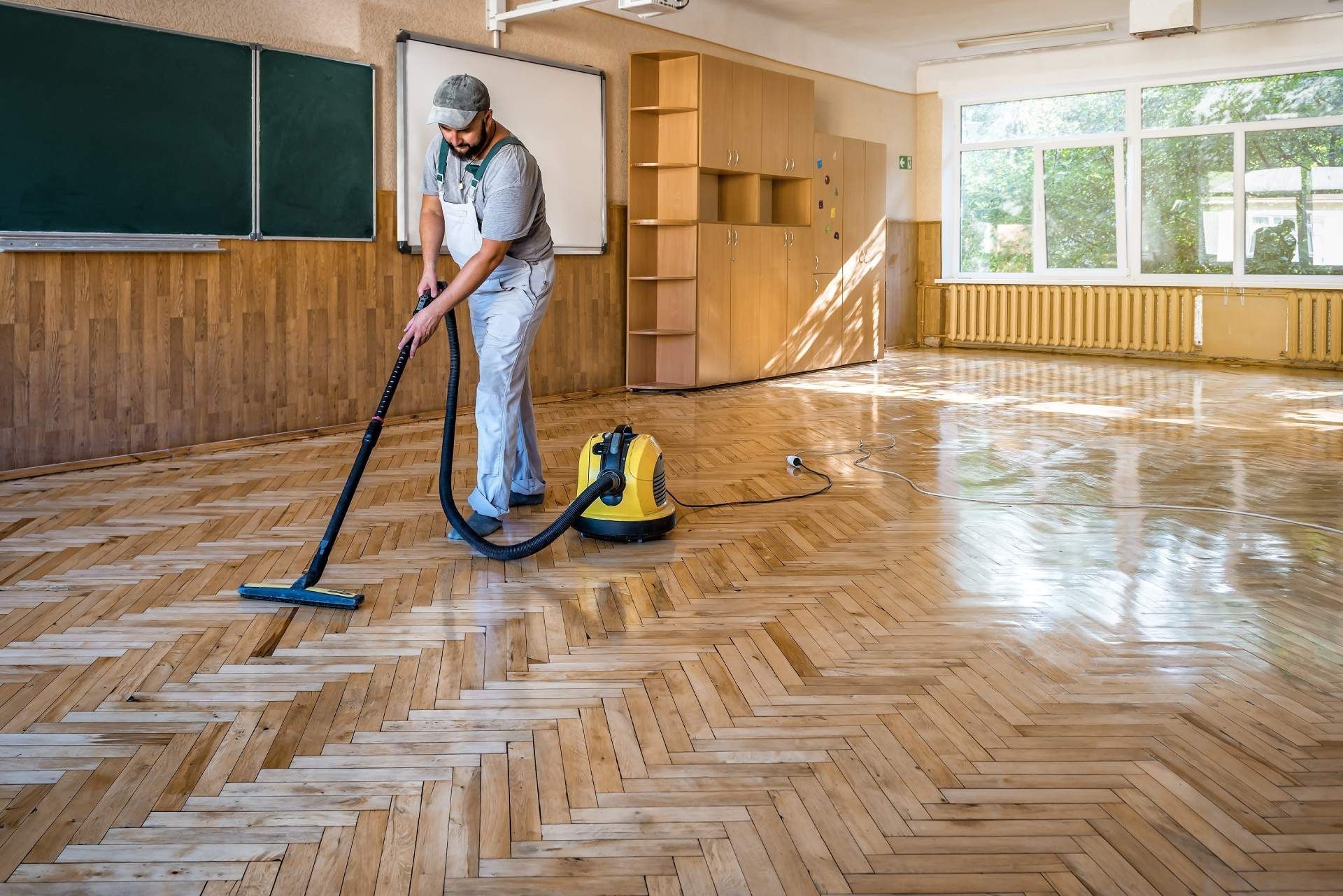 A person in work clothes uses a floor cleaning machine on a polished wooden parquet floor in a school classroom.