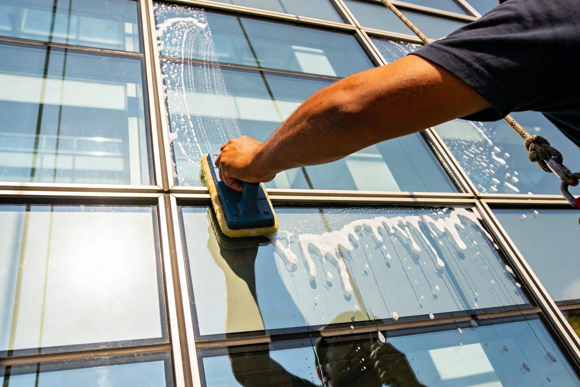 A person uses a handheld scrubber to clean a soapy glass window on the side of a modern building.