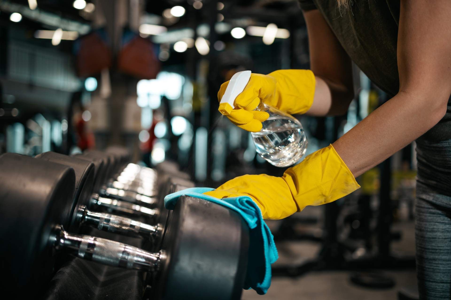 A person wearing yellow rubber gloves cleaning gym dumbbells with a spray bottle and a blue cloth.
