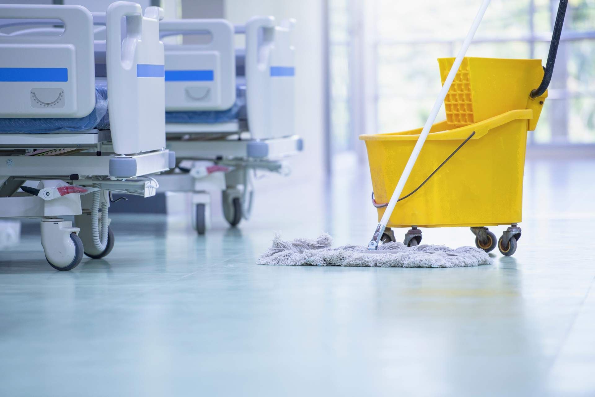 A mop and yellow cleaning bucket on wheels sit on the floor of a brightly lit hospital room near several empty beds.