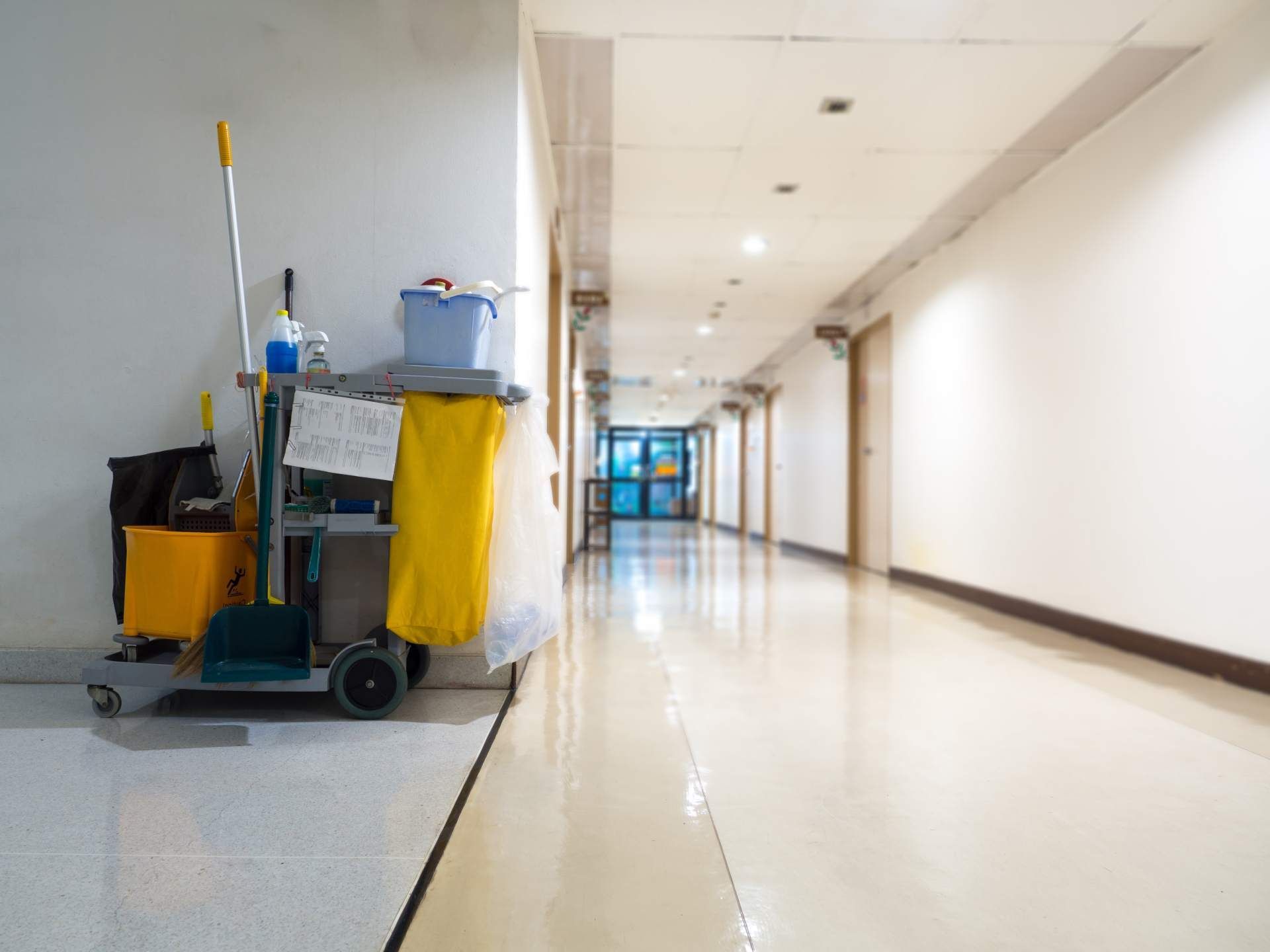 A janitorial cleaning cart sits in the corner of a long, brightly lit building hallway with shiny, polished floors.