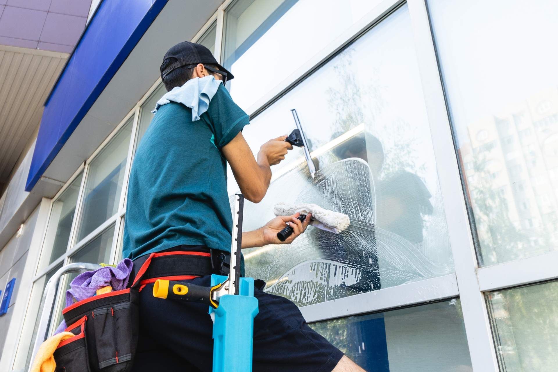 A professional window cleaner wearing a utility belt uses a squeegee and cloth to clean a large storefront window.