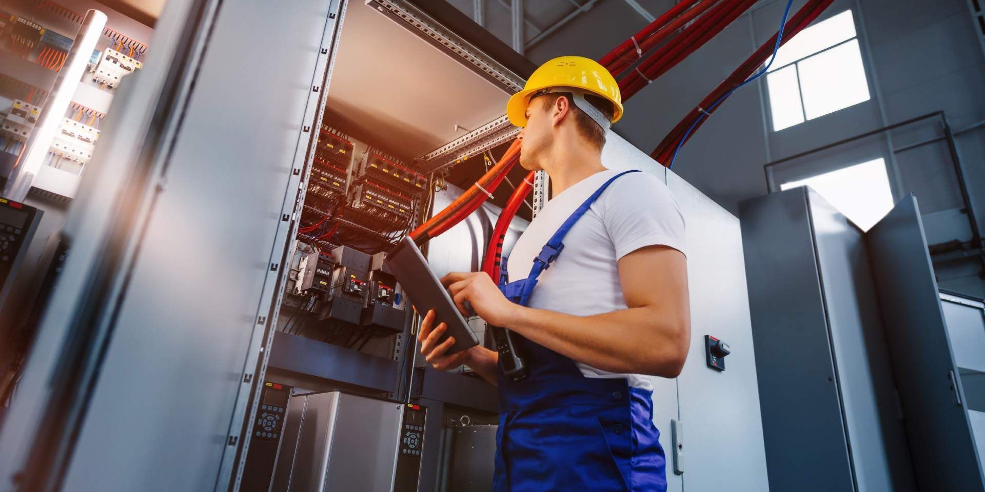 A worker in a yellow hard hat and blue coveralls inspects electrical equipment while holding a tablet.