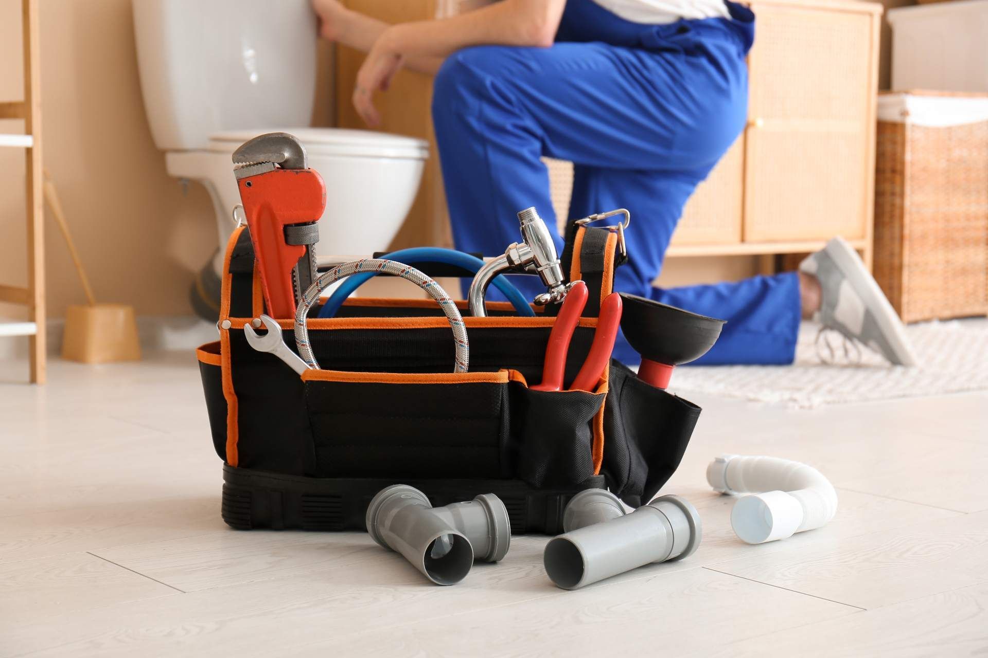 A plumber in blue overalls works on a toilet behind a black tool bag filled with plumbing tools and pipe fittings.