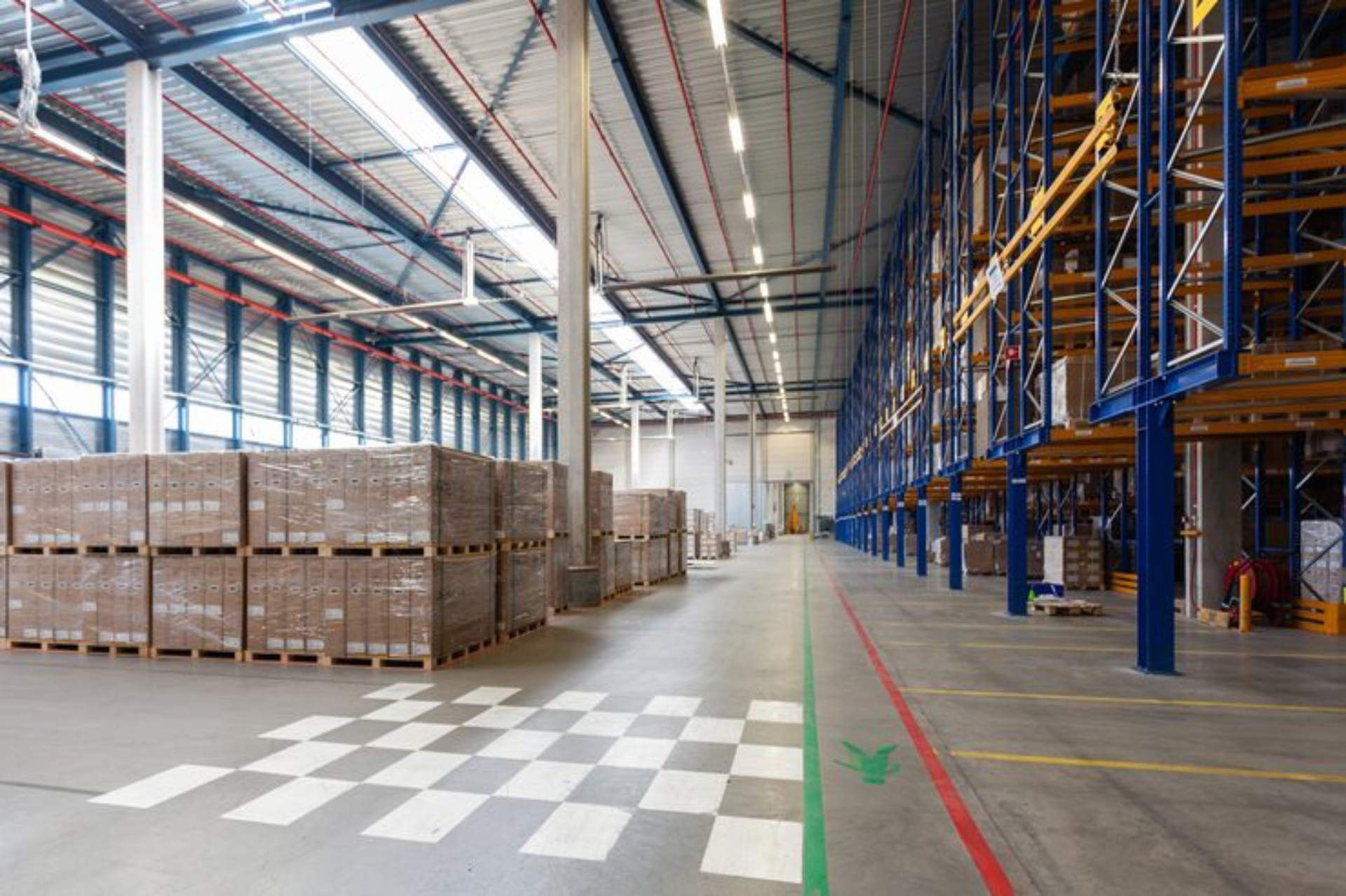 A large, organized warehouse interior featuring tall blue storage racks on the right and stacked pallet boxes on the left.