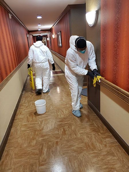Two people in white protective suits and masks clean the walls and floor of a hotel hallway.