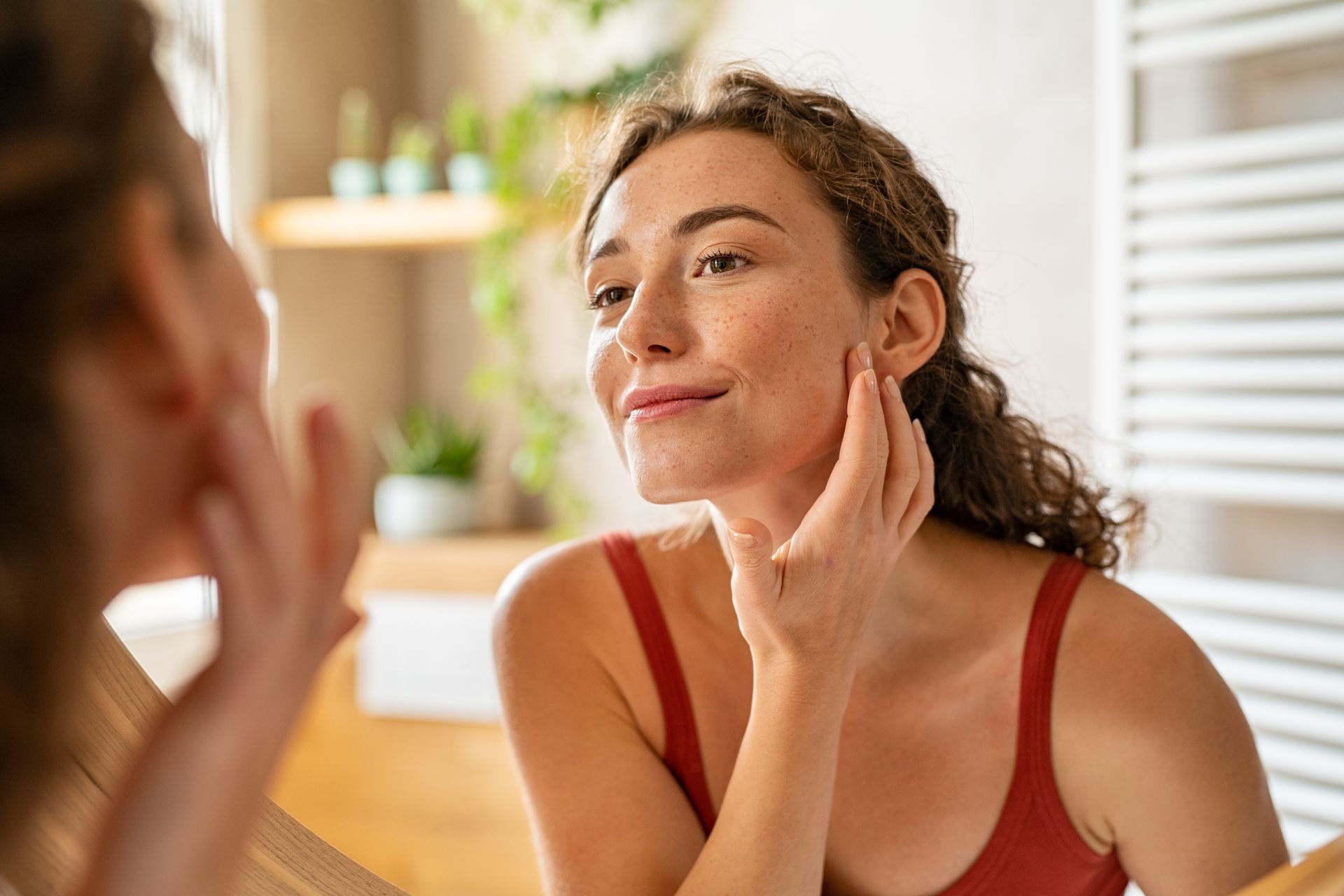 Woman in a red tank top applying skincare to her cheek in a bright bathroom mirror.