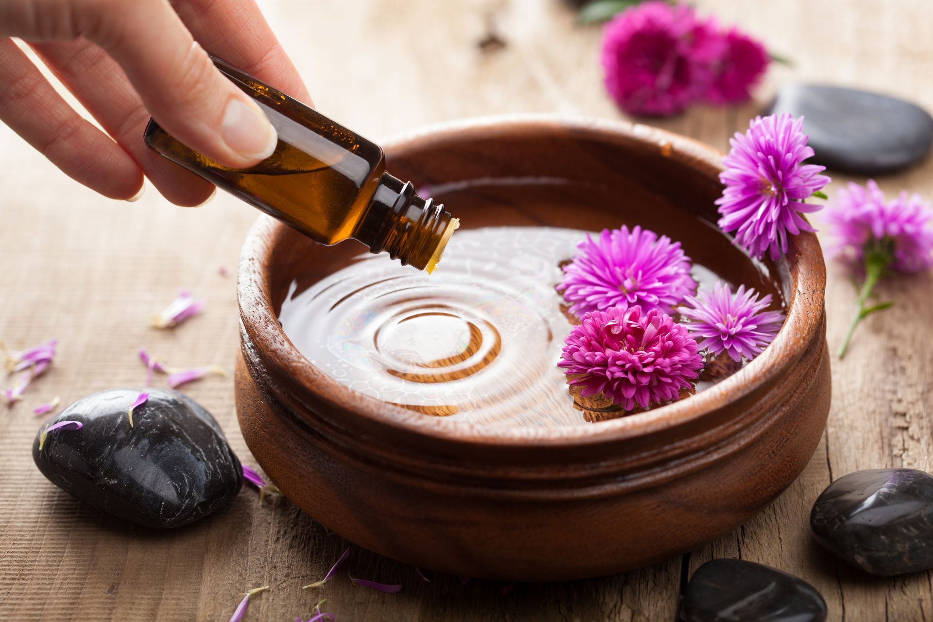 Hand pouring liquid from a bottle into a clay bowl with pink flowers and dark stones on a wooden surface