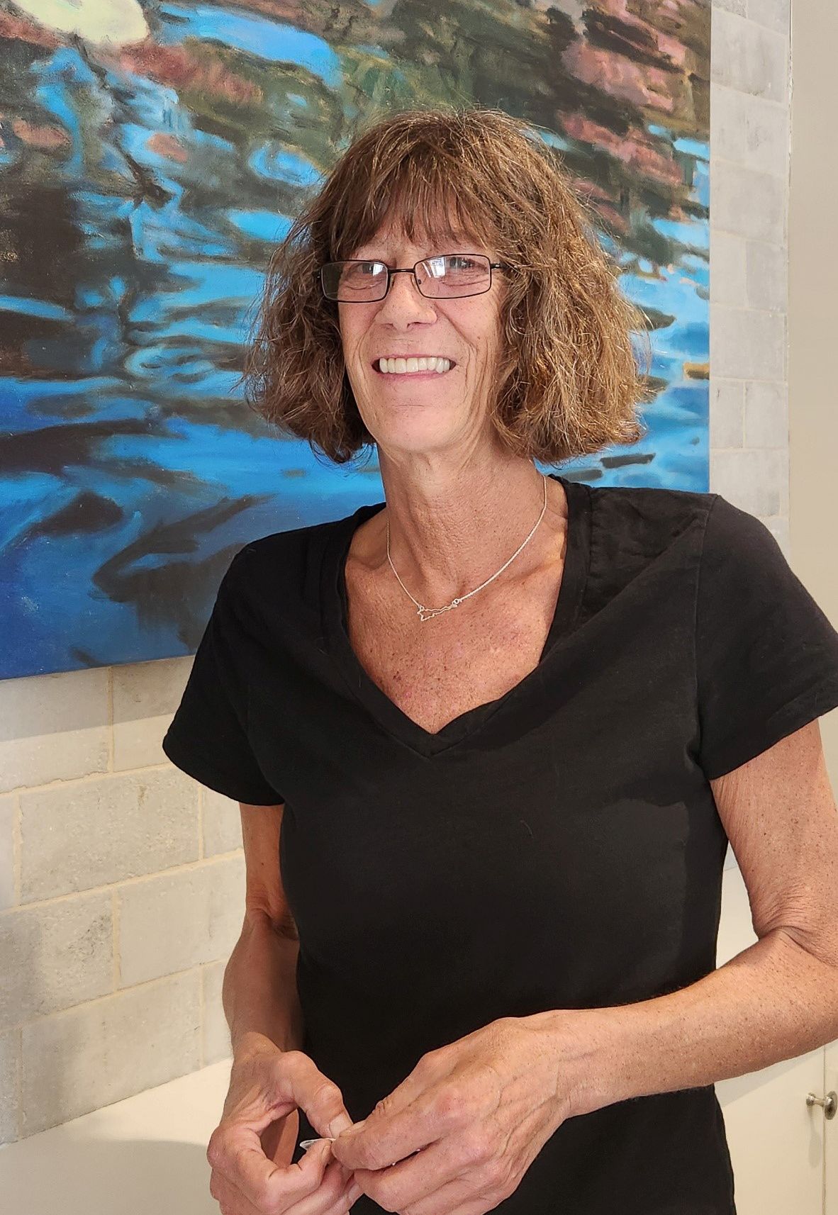 A woman wearing glasses and a black shirt is smiling in front of a painting.